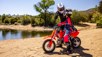 Young rider on bike with instructor on dirt track. Lake in background.
