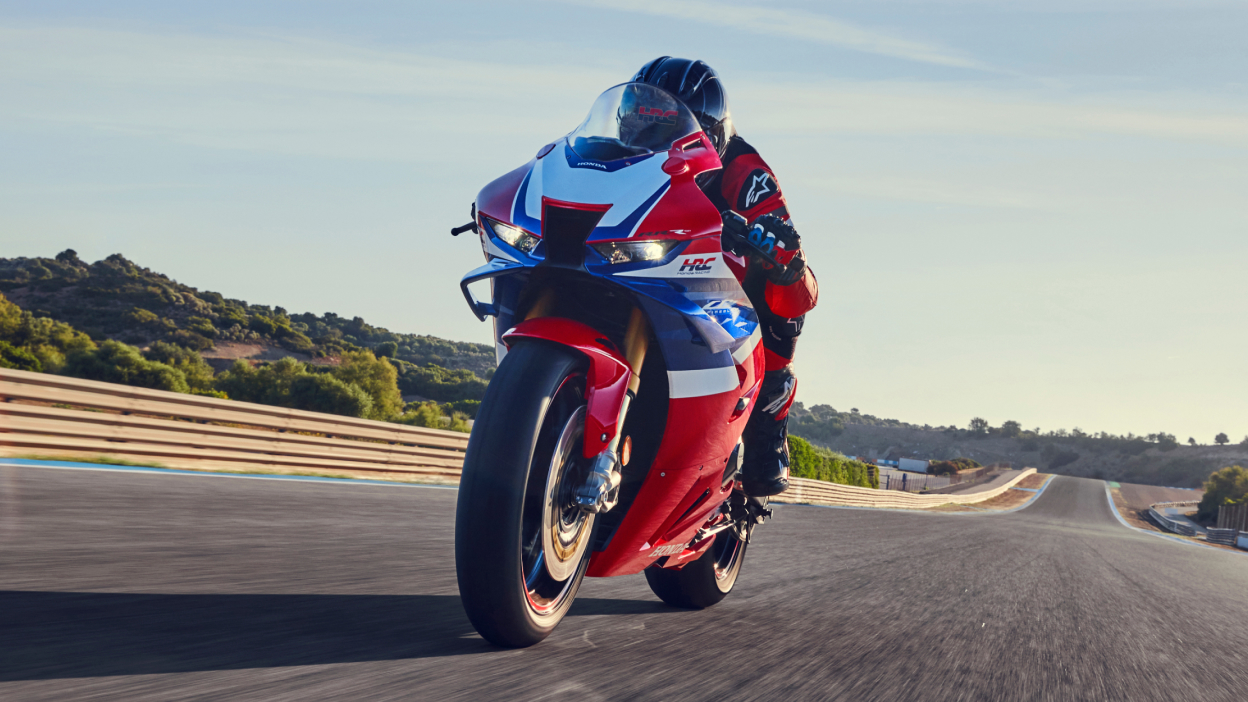 Driver riding Grand Prix Red Fireblade on Track. Front facing. Track in backfroung. Green bushes in behind track background.
