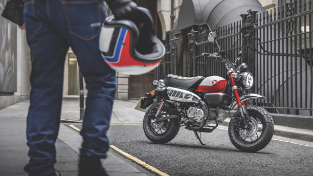 Une personne à partir de la taille, tenant un casque de moto. Une moto est garée devant cette personne. / A person from the waist down, holding a motorcycle helmet. A motorcycle is parked in front of this person.